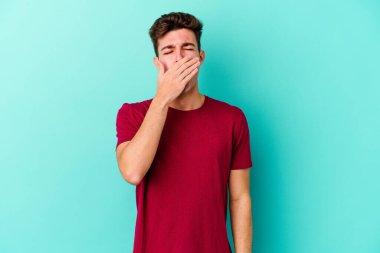 Mavi arka planda izole edilmiş genç beyaz adam esniyor. Ağzı eliyle kapatan yorgun bir hareket sergiliyor..