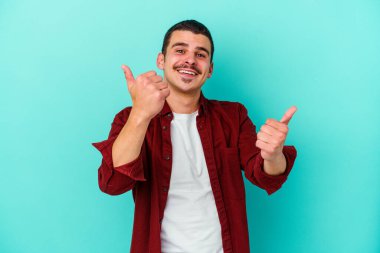 Kafkasyalı genç adam mavi arka planda izole edilmiş. Baş parmağını kaldırıyor, gülümsüyor ve kendinden emin..