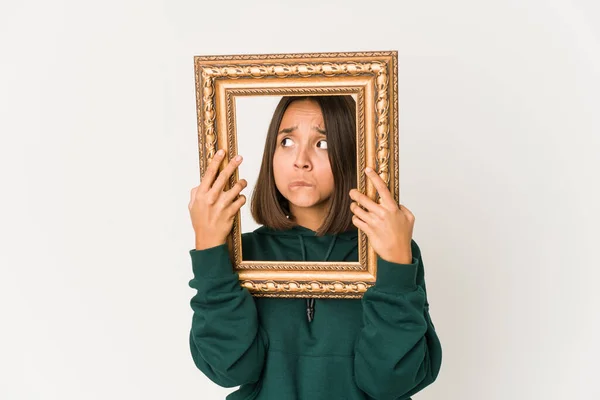Young hispanic woman holding an old frame confused, feels doubtful and unsure.
