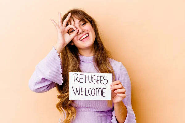 Young caucasian woman holding a Refugees welcome placard isolated excited keeping ok gesture on eye.