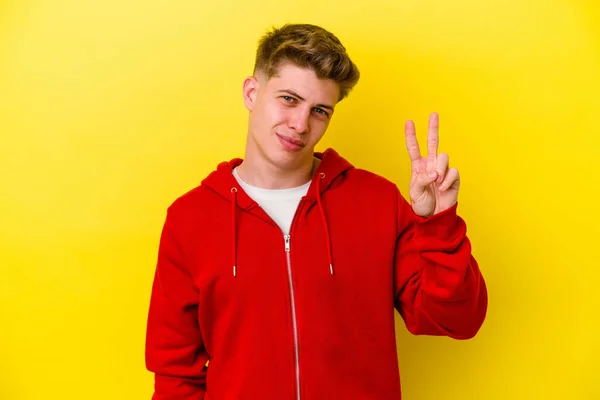 黄色の背景に孤立した若い白人男性喜びと屈託のない指で平和のシンボルを示す — ストック写真