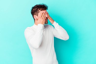 Mavi arka planda izole edilmiş genç beyaz adam elleriyle gözlerini kapatmaktan korkuyor..