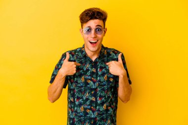 Sarı arka planda izole edilmiş Hawaii tişörtü giyen genç beyaz adam parmağını gösterip gülümseyerek şaşırdı..
