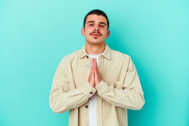 Kafkasyalı genç adam mavi arka planda izole edilmiş dua ediyor, bağlılık gösteriyor, dindar biri ilahi ilham arıyor..