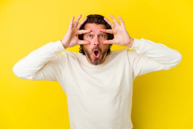 Sarı arka planda izole edilmiş orta yaşlı beyaz bir adam bir başarı fırsatı bulmak için gözlerini açık tutuyor..