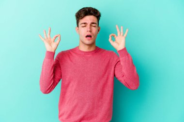 Mavi arka planda izole edilmiş genç beyaz adam yorucu bir günün ardından rahatlıyor. Yoga yapıyor..