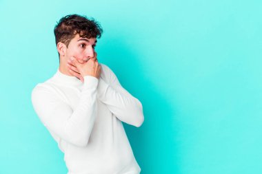 Kafkasyalı genç adam mavi arka planda izole edilmiş. Düşünceli bir şekilde ağzıyla ağzını kapatan bir fotokopi odasına bakıyor..