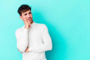 Mavi arka planda izole edilmiş genç beyaz adam şüpheli ve şüpheci bir ifadeyle yana bakıyor..