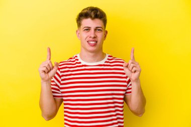 Sarı arka planda izole edilmiş genç beyaz bir adam iki parmağı da boş bir alan gösteriyor..