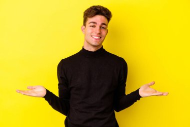 Sarı arka planda izole genç beyaz adam kollarıyla ölçek yapar, mutlu ve kendinden emin hisseder..