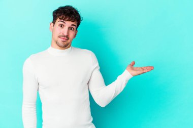 Mavi arka planda izole edilmiş genç beyaz bir adam bir palmiye üzerinde fotokopi alanı ve belinde bir el daha gösteriyor..