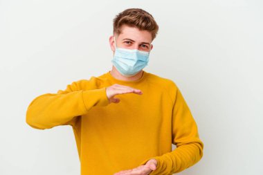Koronavirüs maskesi takan genç adam beyaz arka planda iki eliyle bir şey tutuyor, ürün sunumu..