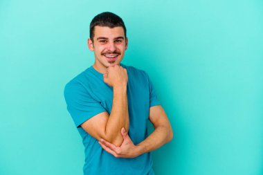 Mavi arka planda izole edilmiş genç beyaz adam gülümsüyor ve kendinden emin, elleriyle çenesine dokunuyor..
