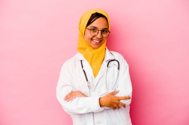 Pembe arka planda izole edilmiş genç bir doktor. Kendine güveni tam, kararlılıkla kollarını kavuşturuyor..