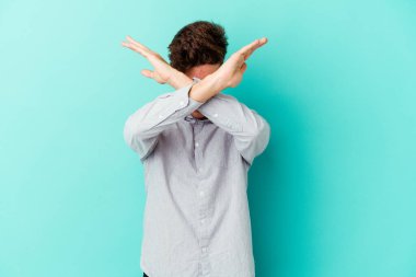 Kafkasyalı genç adam mavi arka planda izole edilmiş iki kolu çapraz tutuyor, inkar kavramı.