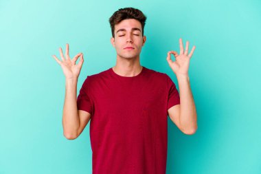 Mavi arka planda izole edilmiş genç beyaz adam yorucu bir günün ardından rahatlıyor. Yoga yapıyor..