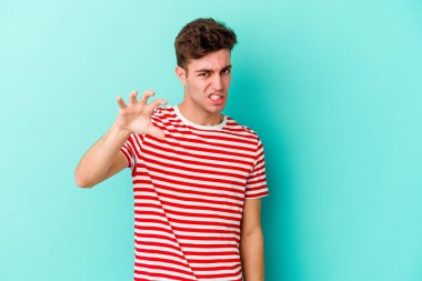 Mavi arka planda izole edilmiş genç beyaz adam pençelerini kedi taklidi yaparken gösteriyor, agresif bir hareket..