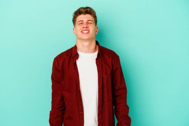 Mavi arka planda izole edilmiş genç beyaz adam gülüyor ve gözlerini kapıyor, rahatlamış ve mutlu hissediyor..