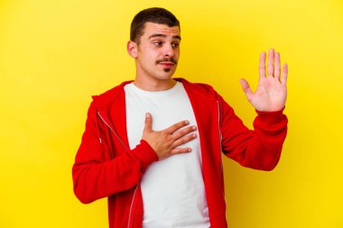 Beyaz tenli genç adam sarı arka planda izole edilmiş yemin ediyor, elini göğsüne koyuyor..