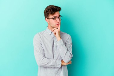 Mavi arka planda izole edilmiş genç beyaz adam alaycı bir ifadeyle kameraya bakarken mutsuz oluyor..