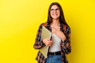 Pembe arka planda dizüstü bilgisayar tutan genç beyaz kadın gülümsüyor ve boş alanda bir şeyler gösteriyor..