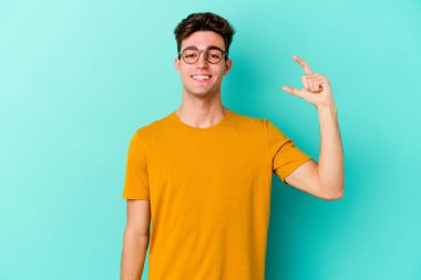 Kafkasyalı genç adam mavi arka planda izole edilmiş. Elinde işaret parmaklarıyla küçük bir şey tutuyor, gülümsüyor ve kendinden emin..