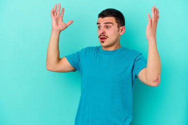 Kafkasyalı genç adam mavi arka planda izole edilmiş gökyüzüne haykırıyor, yukarı bakıyor, hayal kırıklığına uğramış..