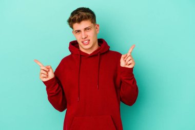 Mavi arka planda izole edilmiş genç beyaz adam farklı kopya alanlarını gösteriyor, birini seçiyor, parmaklarıyla gösteriyor.