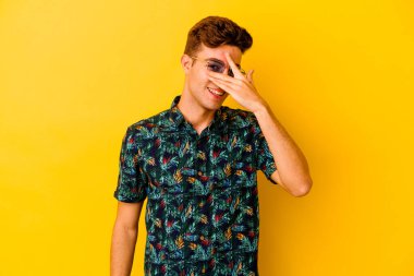 Hawaii gömleği giyen genç beyaz adam sarı arka planda izole edilmiş parmaklarıyla kameraya göz kırpıyor, yüzünü kapatmaktan utanıyor..