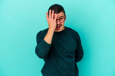 Mavi arka planda izole edilmiş genç beyaz adam yüzünün yarısını palmiye ile kaplayarak eğleniyor..