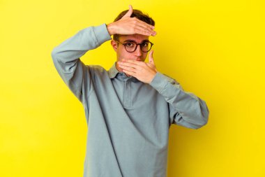 Sarı arka planda izole edilmiş genç beyaz adam kameraya parmaklarıyla göz kırpıyor, yüzünü kapatmaktan utanıyor..