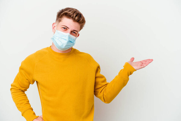 Young man wearing a mask for coronavirus isolated on white background showing a copy space on a palm and holding another hand on waist.