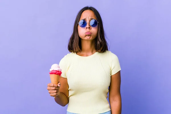 Young mixed race woman eating an ice cream blows cheeks, has tired expression. Facial expression concept.