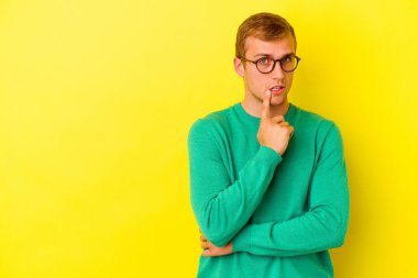 Sarı arka planda izole edilmiş genç beyaz adam şüpheli ve şüpheci bir ifadeyle yana bakıyor..