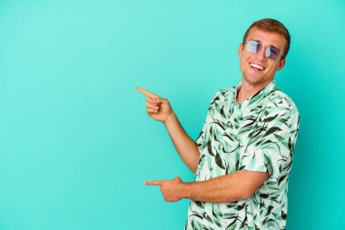 Mavi arka planda izole edilmiş yaz kıyafetleri giyen genç beyaz adam işaret parmaklarıyla işaret ederek heyecanlanıyor..