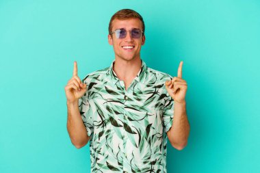 Mavi arka planda izole edilmiş yaz kıyafetleri giyen genç beyaz adam iki ön parmağı da boş bir alan gösteriyor..