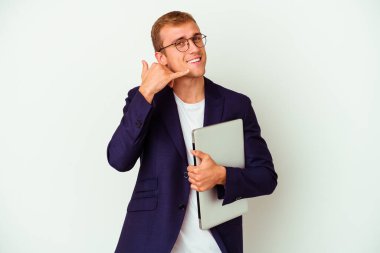 Beyaz arka planda bir dizüstü bilgisayar tutan genç bir beyaz iş adamı parmaklarıyla bir cep telefonu hareketi gösteriyor..