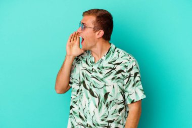 Mavi arka planda izole edilmiş yazlık giysiler giyen genç beyaz bir adam bağırıyor ve ağzı açık bir şekilde avuç içi tutuyor..