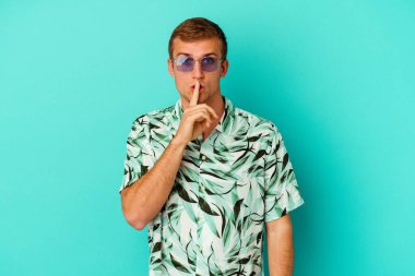Mavi arka planda izole edilmiş yaz kıyafetleri giyen genç beyaz bir adam bir sır saklıyor ya da sessizlik istiyor..