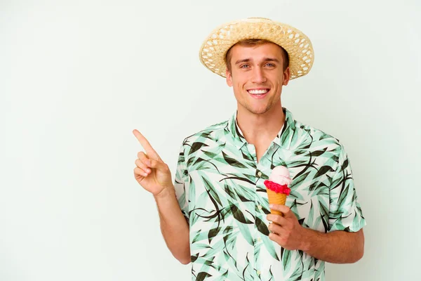 夏の服を着た若い白人の白人男性が 白い背景の上に孤立したアイスクリームを持ち 笑顔と脇を指差して 空白のスペースで何かを示しています — ストック写真