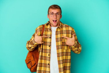 Kafkasyalı genç bir öğrenci mavi arka planda izole edildi. Parmakla göstererek şaşırdı, gülümsedi..