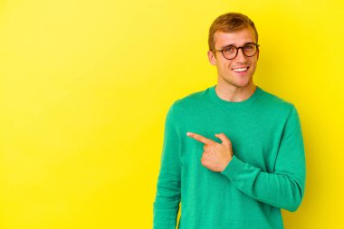 Sarı arka planda izole edilmiş genç beyaz adam gülümsüyor ve boş alanda bir şeyler gösteriyor..