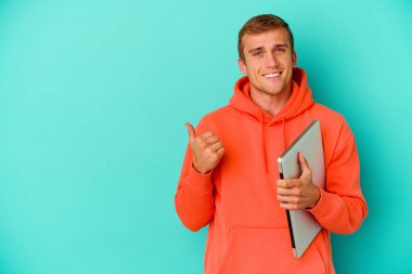 Genç beyaz bir adam dizüstü bilgisayar tutuyor mavi arka planda izole edilmiş gülümsüyor ve başparmağını kaldırıyor.