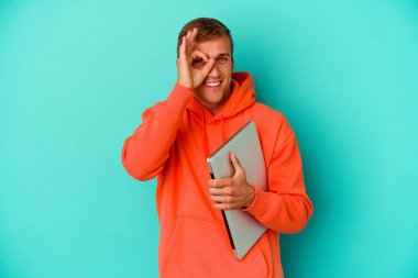 Mavi arka planda dizüstü bilgisayar tutan genç beyaz bir adam heyecanlı. Gözünü üzerinden ayırma..