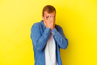 Sarı arka planda izole edilmiş genç beyaz adam parmaklarının arasından korkmuş ve gergin bir şekilde göz kırp..