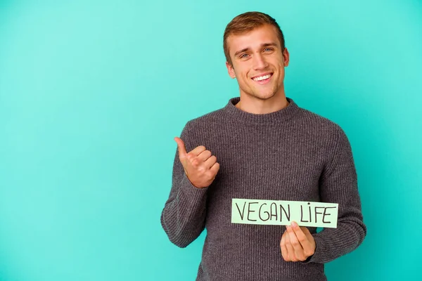 年轻的高加索人拿着素食主义者的生活标语牌 被蓝色背景隔离 微笑着举起大拇指 — 图库照片