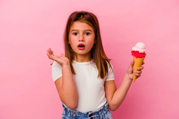 拿着粉红色背景的冰淇淋的高加索小女孩感到惊讶和震惊 — 图库照片