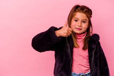 Pembe arka planda izole edilmiş küçük beyaz kız gülümsüyor ve başparmağını kaldırıyor.