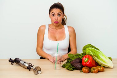 Genç beyaz kadın sebzeli sağlıklı bir smoothie hazırlıyor omuzlarını silkiyor ve gözleri açık..