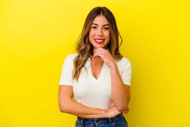 Sarı arka planda izole edilmiş genç beyaz kadın gülümsüyor ve kendinden emin, elleriyle çenesine dokunuyor..